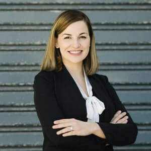 A woman with straight, shoulder-length hair in a black blazer and white blouse stands in front of gray metal steps, smiling with arms crossed.