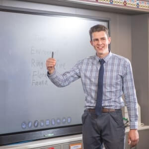 A man in a checkered shirt and tie stands in front of a smartboard, holding a stylus and smiling, with words written on the board behind him.