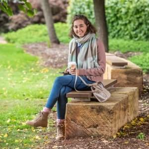 A woman sits on a wooden bench in a park, holding a takeaway coffee cup. She is wearing a scarf, jacket, jeans, and boots, with a small bag next to her.