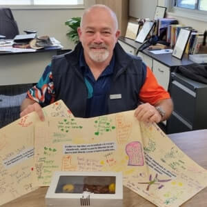 A smiling man sits at a desk holding several large, handwritten cards and a box of assorted pastries in an office setting.