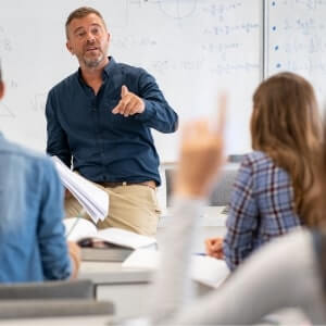 A male teacher sits on a desk in front of a whiteboard, addressing students; one student in the foreground has their hand raised.