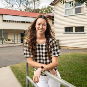 A woman with long brown hair, wearing a checkered shirt and white pants, leans on a metal railing outside a cream-colored building with a red roof.