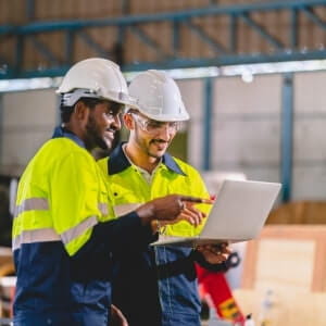 Two workers in safety helmets and high-visibility jackets look at a laptop and discuss something inside an industrial warehouse.