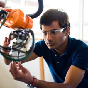 A man wearing safety glasses inspects or adjusts a piece of industrial machinery or robotic equipment indoors.