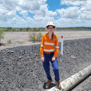 A person in a high-visibility orange and blue work uniform and hat stands on gravel near a pipeline in an open, outdoor area under a partly cloudy sky.