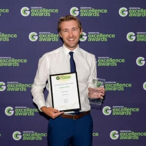 A man in a white shirt and blue tie holds a certificate and a glass trophy in front of a backdrop reading "Geospatial Excellence Awards.