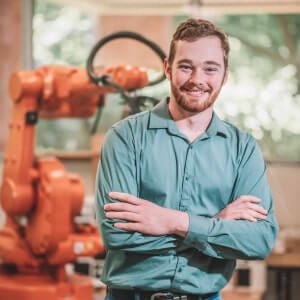 A man in a teal shirt stands with arms crossed, smiling in front of an orange industrial robot in a bright workspace.