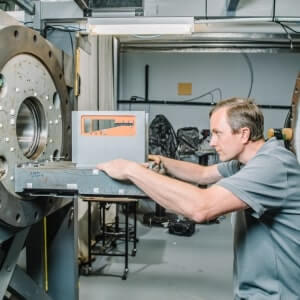 A man in a gray shirt operates industrial machinery in a workshop, adjusting a control panel beside a large metal component.