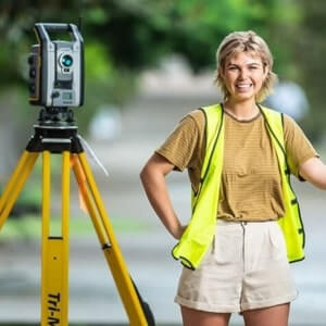 Person wearing a yellow safety vest stands next to a surveying instrument mounted on a tripod outdoors.