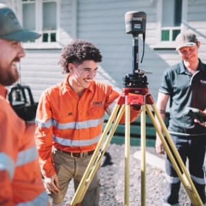 Three people in high-visibility work clothes stand around a surveying tripod with equipment outside a building.
