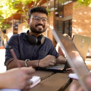 A person with glasses and headphones around their neck is smiling while using a laptop at an outdoor table with others.