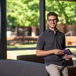 A man wearing glasses and a dark polo shirt stands indoors, smiling and holding a book, with large windows and greenery visible in the background.