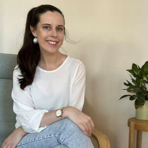 Woman with long dark hair in a ponytail, wearing a white blouse and jeans, sits on a light grey chair next to a small table with a potted plant.