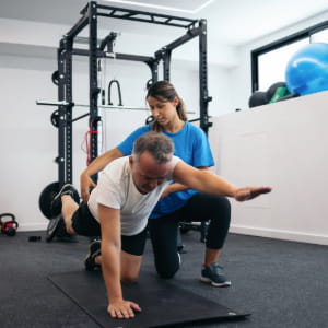 A woman in a blue shirt providing support for a man doing exercises on the floor.