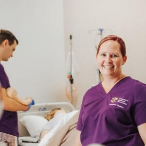 A smiling healthcare worker in purple scrubs stands in the foreground, while another tends to a patient in a hospital bed in the background.
