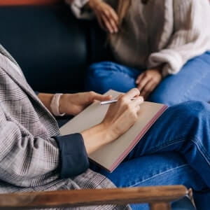 Two people sit together; one holds a notebook and pen, appearing to take notes or write while the other listens.