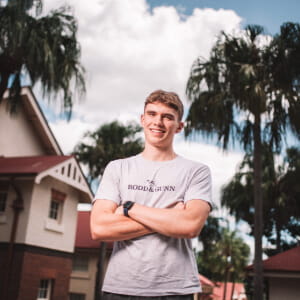 Young man in a light t-shirt stands outdoors with arms crossed, smiling, in front of palm trees and buildings under a partly cloudy sky.