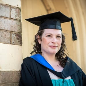 A person wearing a graduation cap and gown stands against a brick wall, looking at the camera with a slight smile.