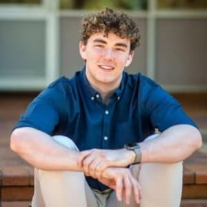 A young man with curly hair, wearing a dark blue shirt and light pants, sits on outdoor steps, smiling at the camera.