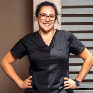 Person wearing black scrubs and glasses stands with hands on hips, smiling, in front of a modern background with horizontal slats.