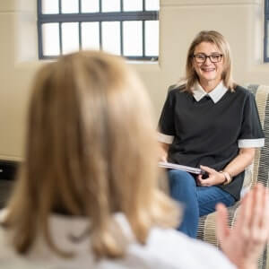 Two women sit facing each other in conversation; one is smiling and holding a notebook, suggesting a counseling or interview session.