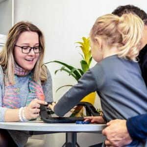 A woman and a man assist a young girl using a tablet at a small table, with a plant in the background.