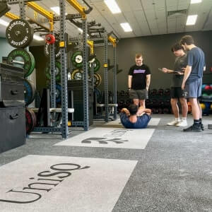 Four people are in a gym; one is doing sit-ups on the floor while the others stand nearby, observing and holding a clipboard. Weightlifting equipment is visible in the background.