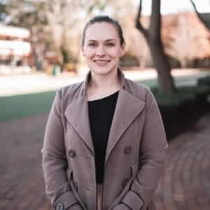 A woman with light skin and pulled-back hair stands outdoors on a brick path, wearing a taupe coat over a black top, with trees and buildings in the background.
