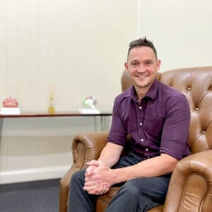 A man wearing a purple shirt sits on a brown leather chair, smiling. A table with decorative items is visible in the background against a cream wall.