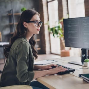 A woman wearing glasses types on a keyboard while looking at code on a computer monitor in a modern office setting.