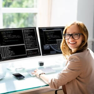 A woman wearing glasses sits at a desk with two monitors displaying lines of code, smiling toward the camera in a bright office setting.