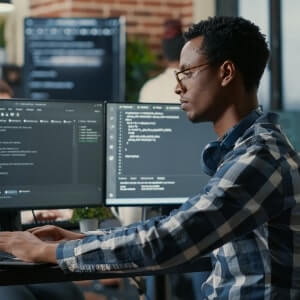 A person wearing glasses and a plaid shirt works at a desk with dual monitors displaying code in a modern office setting.