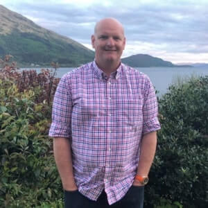 A man in a pink checkered shirt stands outdoors with his hands in his pockets, with greenery, a body of water, and hills in the background.