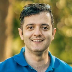 A man with short dark hair, wearing a blue collared shirt and sunglasses on his head, smiles outdoors with a blurred green background.