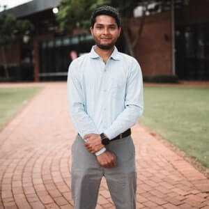 A man in a light blue dress shirt and gray pants stands on a brick pathway with green grass and a brick building in the background.