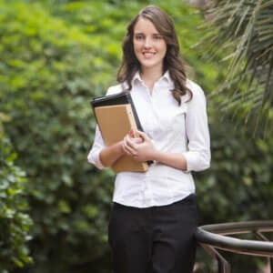A woman in a white shirt and black pants stands outdoors, holding a folder and smiling, with greenery in the background.