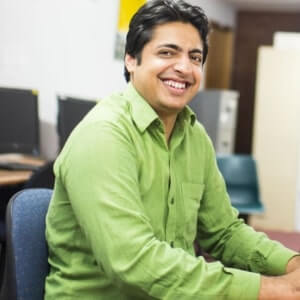 A man in a green shirt sits on an office chair, smiling, with computers and office furniture in the background.