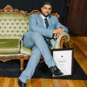 A man in a light blue suit sits on a vintage green sofa, holding a framed certificate with his arm resting on the armrest, on a wooden floor background.