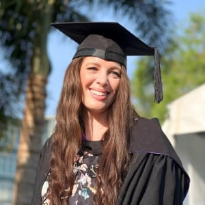 A woman wearing a graduation cap and gown stands outdoors, smiling, with trees and buildings in the background.