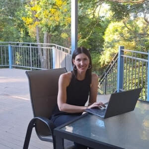 A woman in black sits at a glass table outdoors, working on a laptop, with trees and a metal railing in the background.