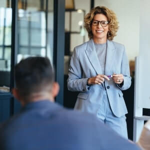 A woman in a gray suit stands and smiles while holding a marker, speaking to a seated person in an office setting.