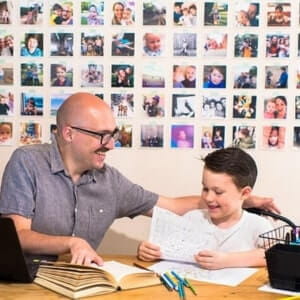An adult and a child sit at a table with books and papers, smiling, in front of a wall covered with small family photos.
