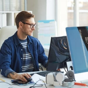 A man wearing glasses and a striped shirt works at a desk with a computer monitor and office supplies in a bright, modern office setting.