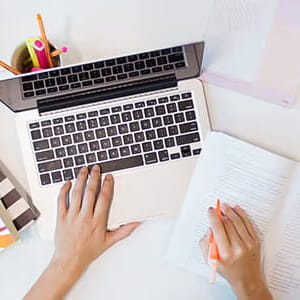 A person writes in a notebook with one hand while using a laptop with the other, surrounded by stationery items on a white desk.