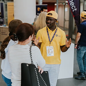 A man in a yellow polo shirt and cap speaks to two women indoors during an event, wearing an ID badge and lanyard. Other people are visible in the background.