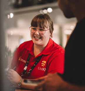 A person wearing a red UniSQ shirt and glasses smiles while talking to another person indoors.