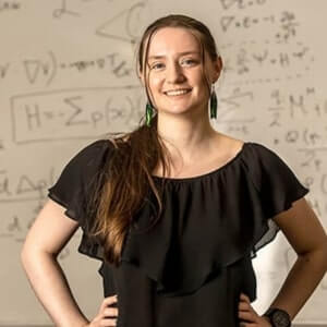 A woman in a black blouse stands in front of a whiteboard covered with mathematical equations, smiling at the camera with her hands on her hips.
