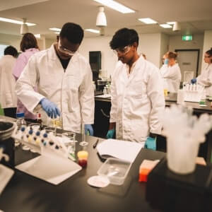 Two people in lab coats and gloves work with scientific equipment in a laboratory setting, with others working in the background.