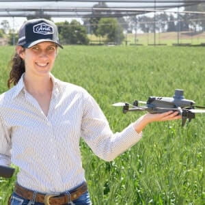 A woman standing in a field holds a drone in one hand and a remote control in the other, smiling at the camera.