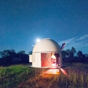 A person stands at the entrance of a small observatory dome at night, with stars visible in the clear sky above.